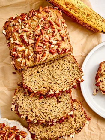 A loaf of protein banana nut bread studded with chopped pecans on a cutting board, with a few slices in front of the loaf and another slice on a plate nearby. Ripe bananas in the background.