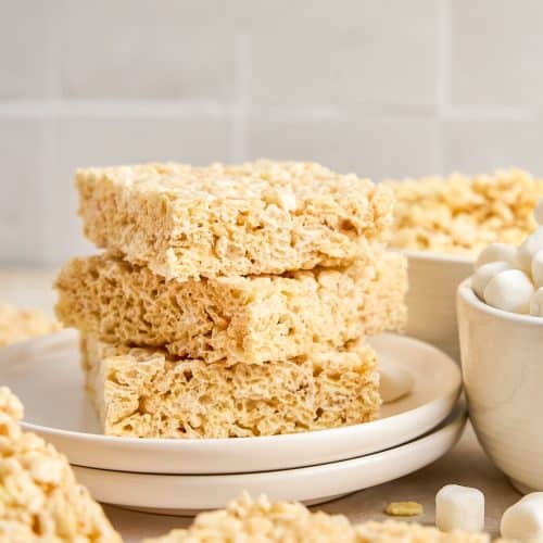 A stack of three rice krispie treats on a plate with more treats and mini marshmallows nearby.